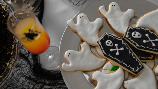 Des gateaux en forme de fantômes et de cercueils dans une assiette blanche un verre contenant un jus orange, le tout posé sur une table avec une nappe blanche.